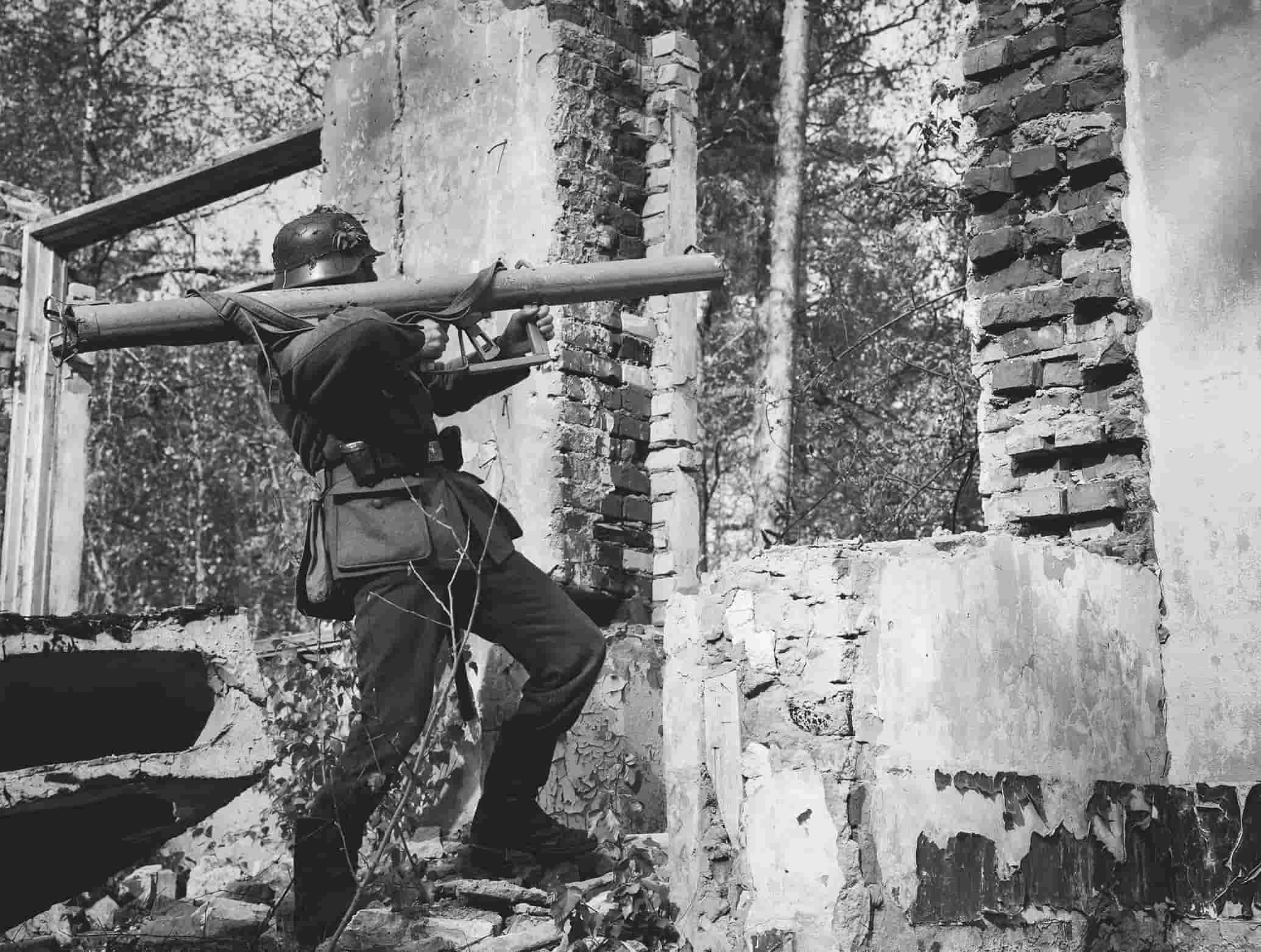 A soldier in uniform stands among ruined brick walls, aiming a long weapon over his shoulder in an outdoor setting. The image is in black and white.